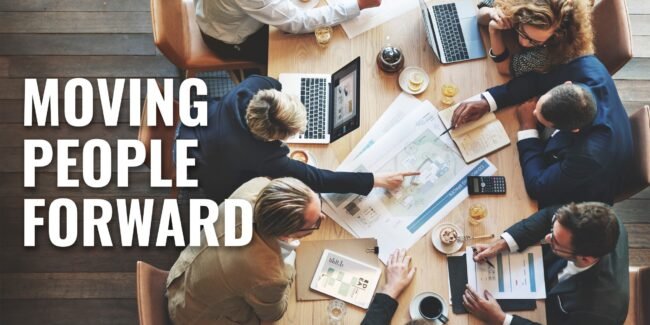 A group of professionals working at a table with a mix of computers and paperwork. The words moving people forwards on the side.